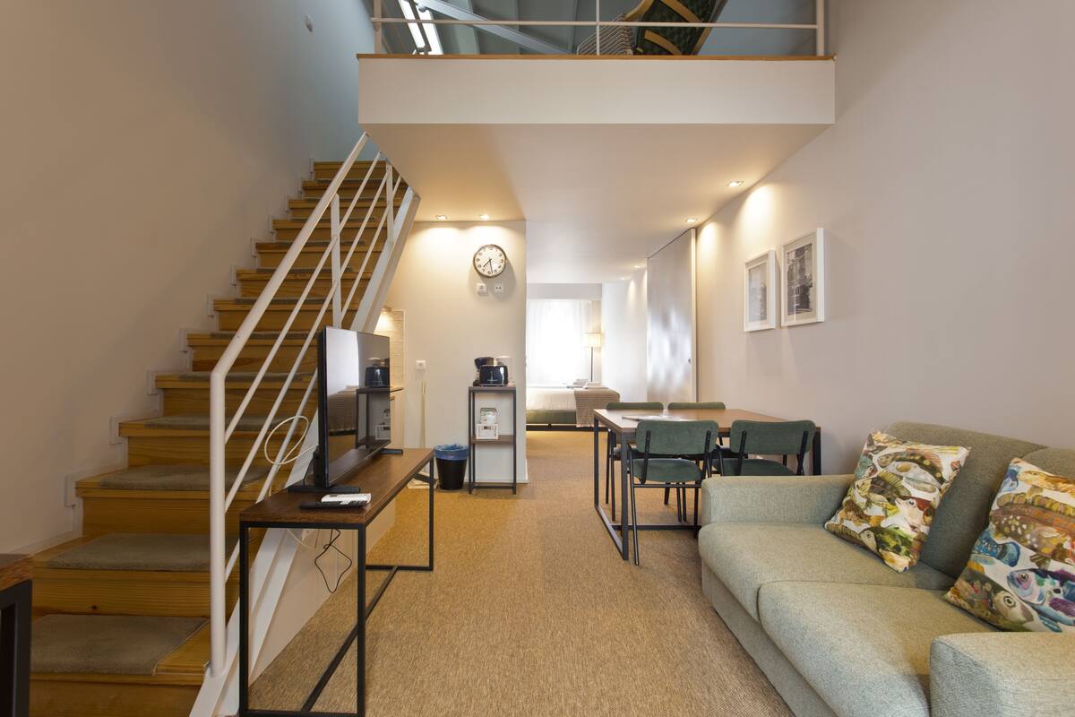 A modern living room with a staircase, a dining area, and a sofa with patterned cushions.