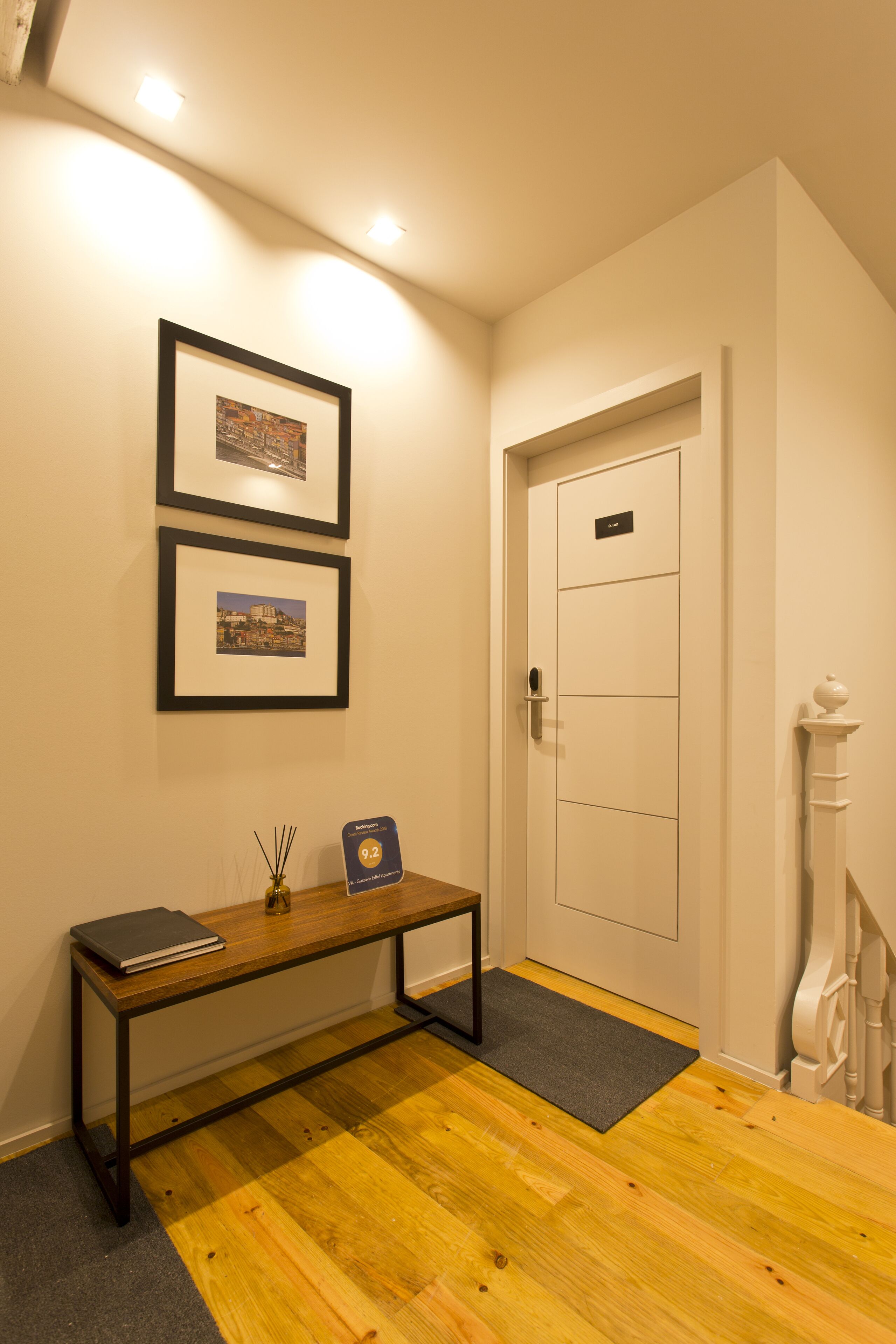 A hallway with a wooden bench, two framed pictures on the wall, a door with a nameplate, and a white staircase.