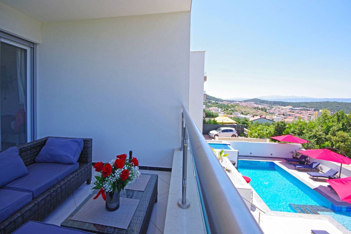 A balcony with a wicker sofa, a table with a vase of flowers, and a pool area with lounge chairs and umbrellas.