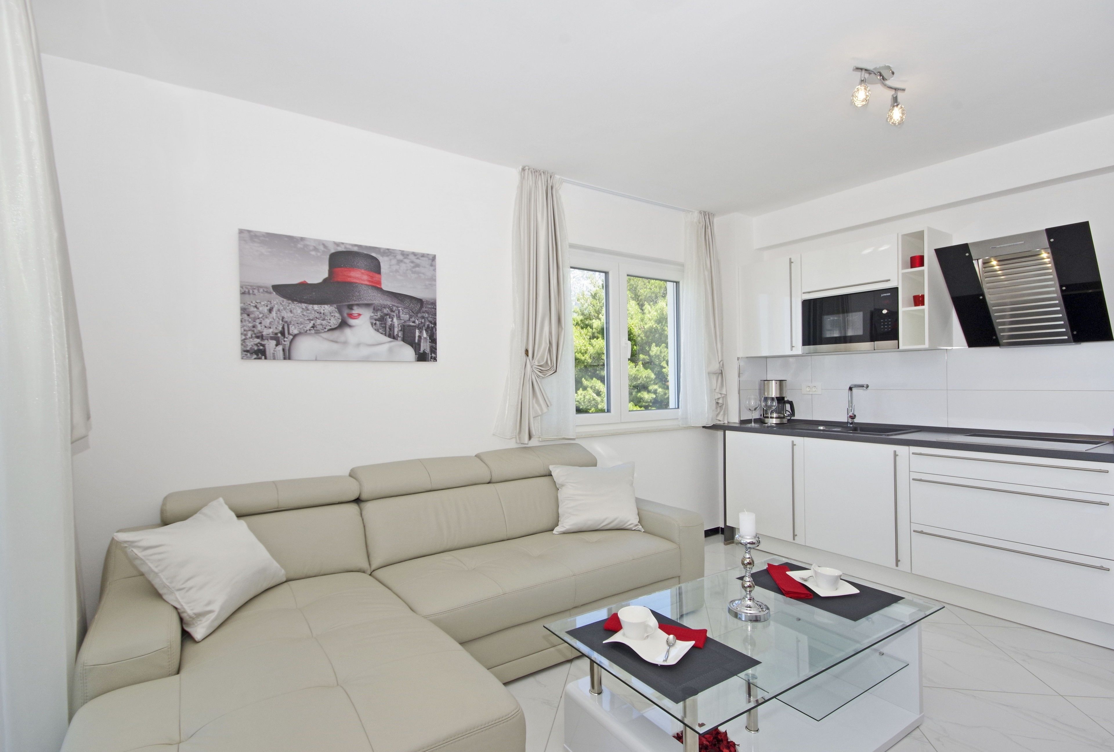 Una sala de estar moderna con un sofá beige, una mesa de centro de vidrio y una zona de cocina con armarios blancos y un microondas.