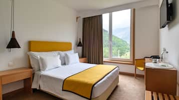 A hotel room with a bed, a bedside table, a desk, a chair, a window with a view of greenery, and a mounted TV.