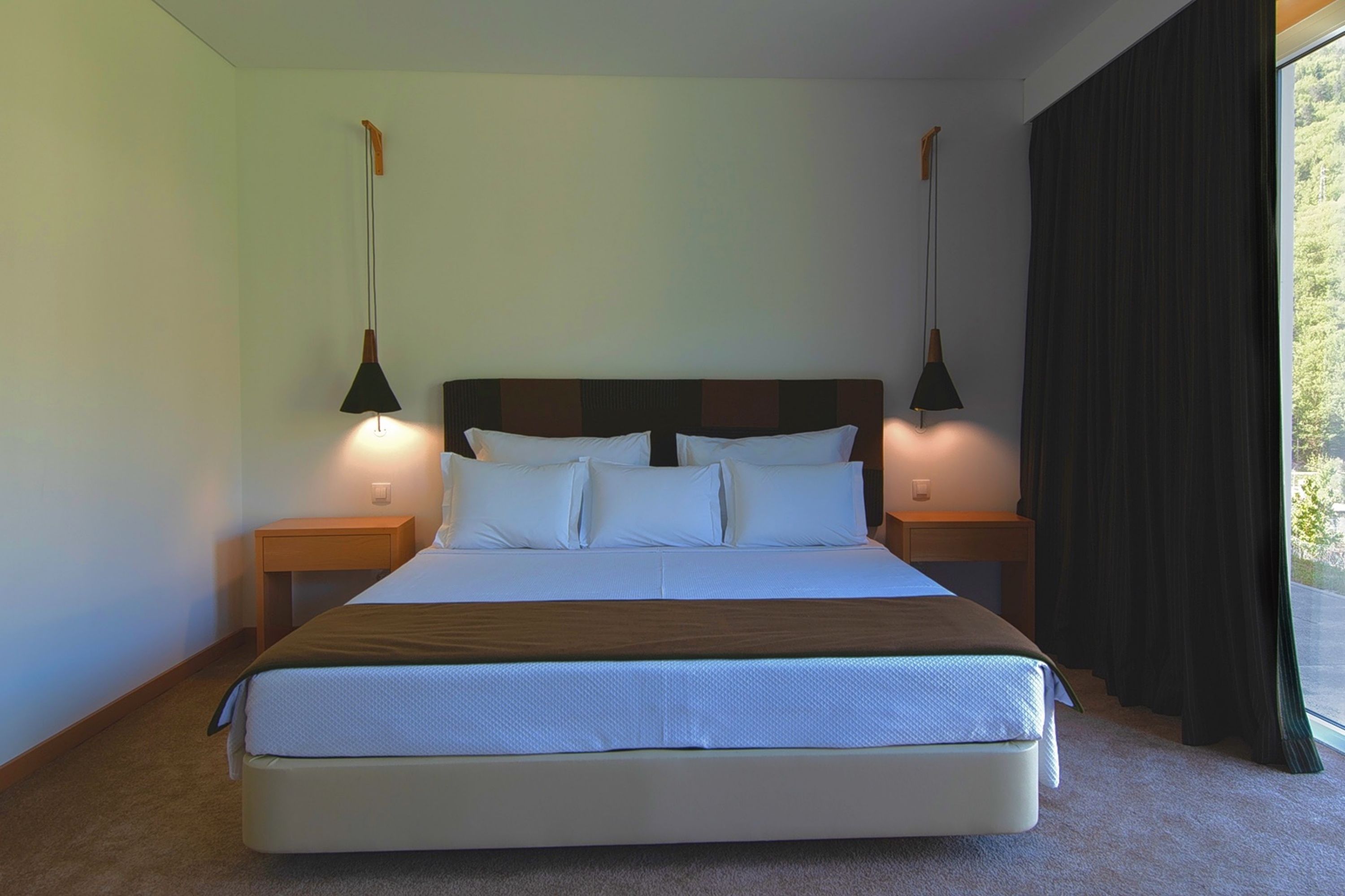 Una cama bien hecha con cabecera de madera, dos mesitas de noche con lámparas, y vistas a la vegetación a través de una ventana con cortinas oscuras.