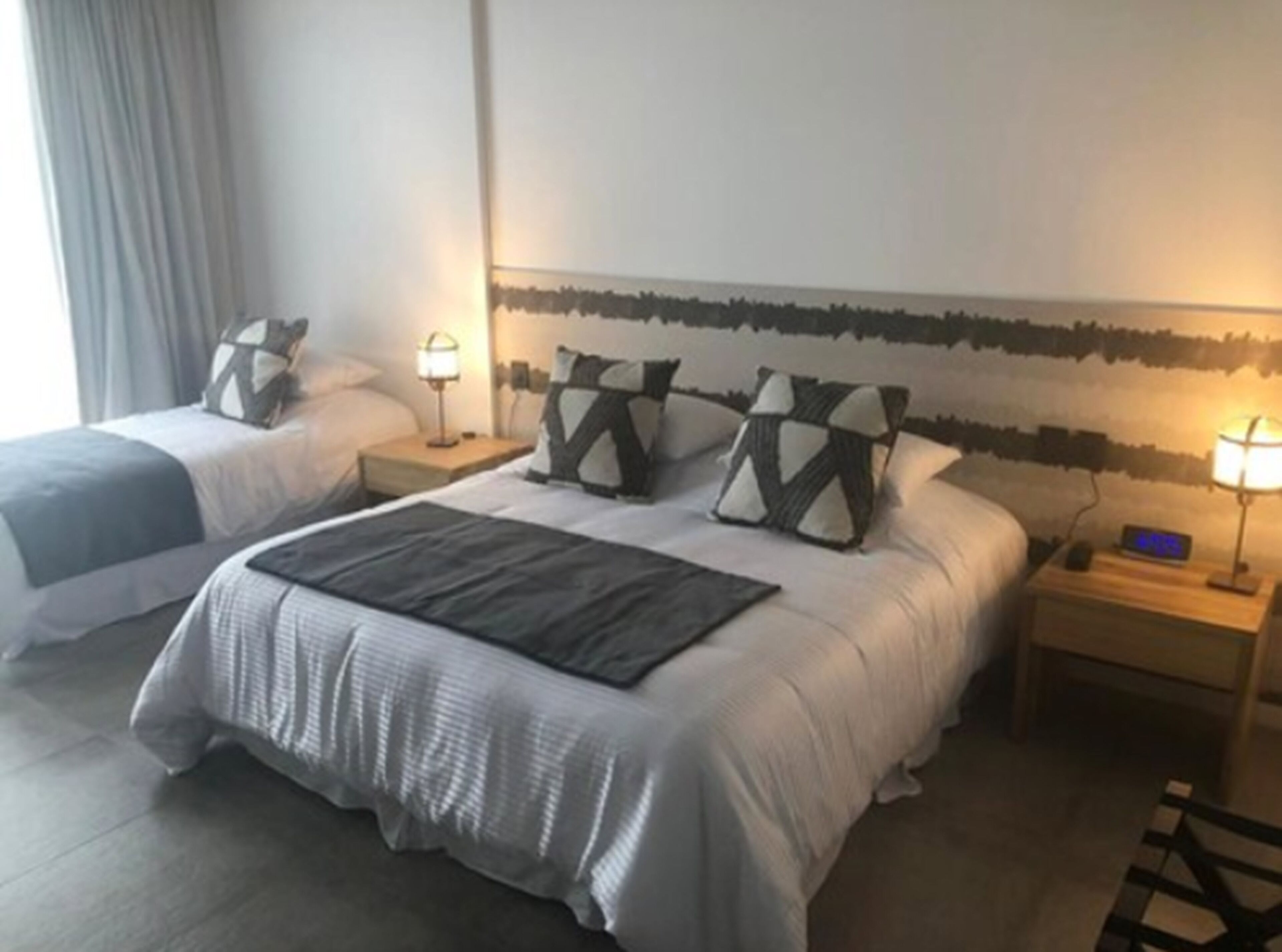 Habitación de hotel con dos camas, cada una con mesita de noche, lámpara y reloj digital.