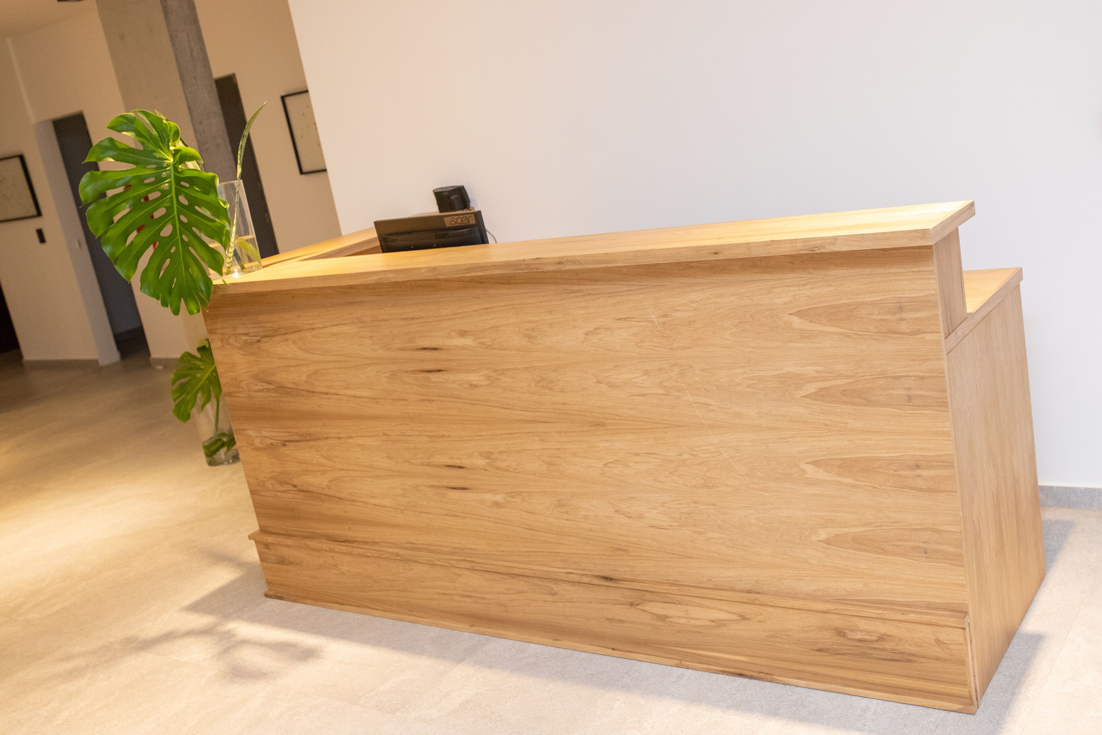 A wooden reception desk with a glass top, positioned in a room with a light-colored floor and a large green plant to the left.