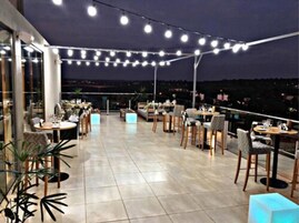 Un área de comedor al aire libre con luces colgantes, mesas, sillas y plantas en macetas.