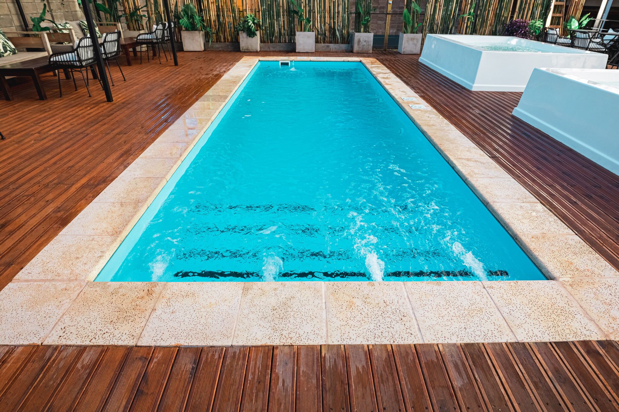 A rectangular indoor pool with a wooden deck surrounding it, featuring a few chairs and tables.