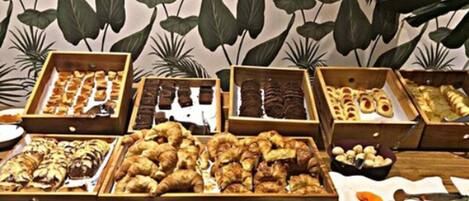 Variedad de productos horneados expuestos en cajas de madera, incluyendo pasteles y pan, sobre una mesa con un fondo de hojas verdes grandes.