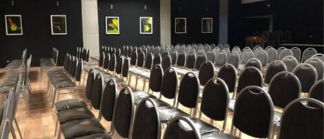 Ein Konferenzraum mit Reihen schwarzer Stühle und Tische, schwarzen Wänden mit gerahmten Bildern und einer Bühne.