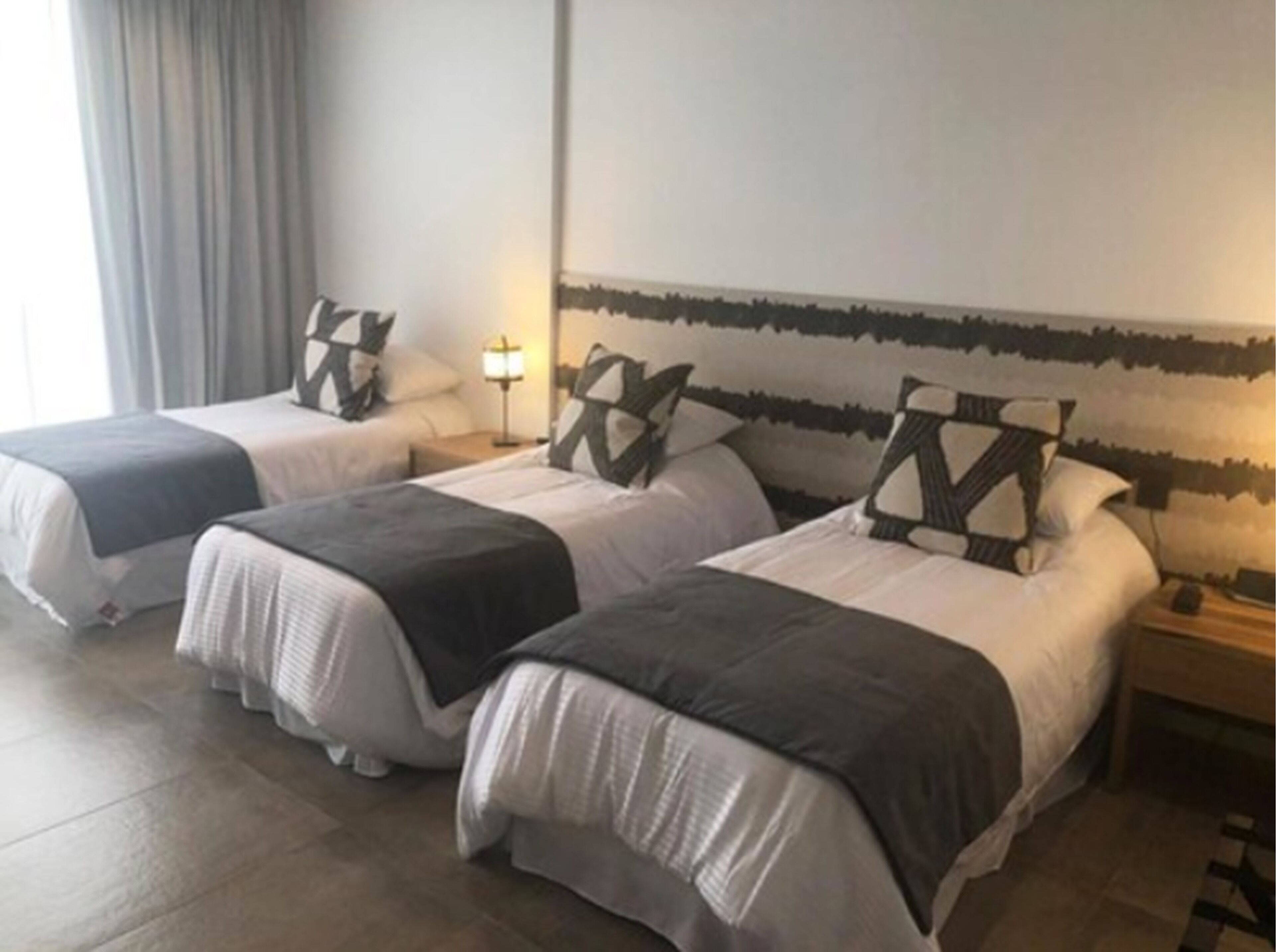 A hotel room with three beds, each with a black and white patterned headboard, a bedside table with a lamp, and a large window with curtains.