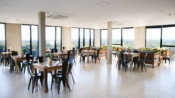 Un comedor amplio con varias mesas y sillas, ventanas grandes que ofrecen vistas a la ciudad y un techo alto con vigas a la vista.