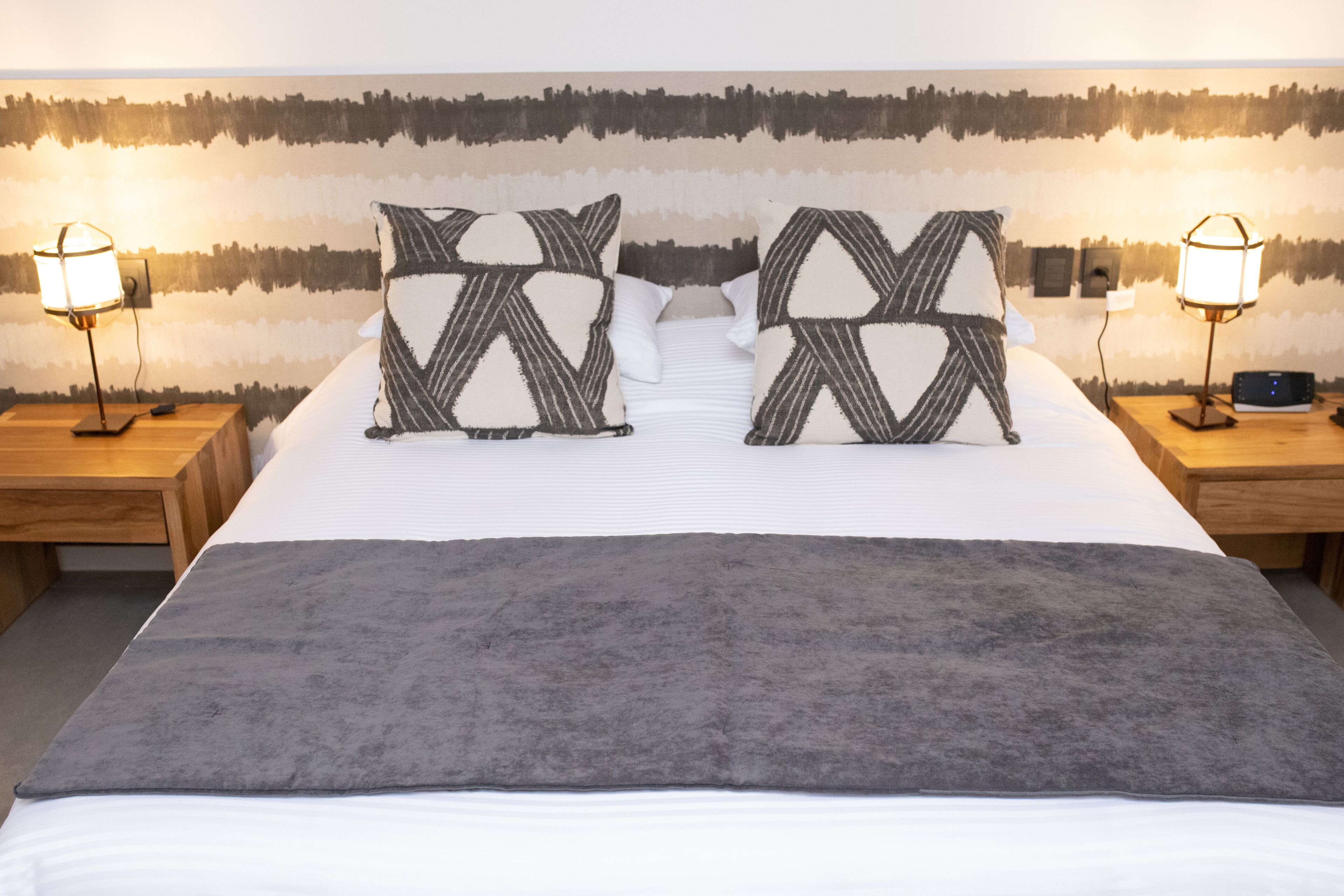 A neatly made bed with two patterned pillows, a gray throw blanket, and two bedside lamps on wooden nightstands.
