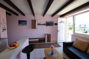 Sala de estar com sofá, mesa de centro, televisão, área de cozinha com frutas na bancada e uma porta que dá para o exterior.