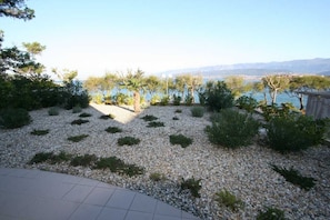 A landscaped garden with gravel paths, various shrubs, and a view of a body of water and distant mountains. - Apartment Djakovic Boba nr 1 (Silo - island Krk)