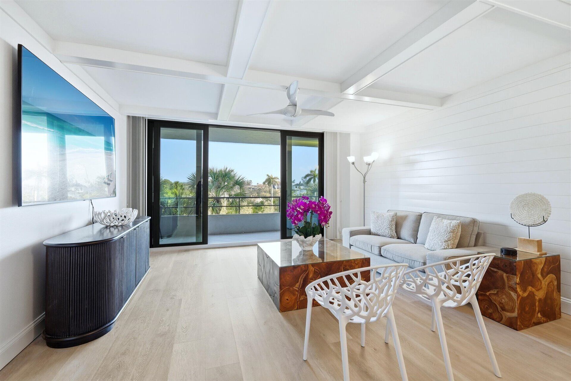 Un salon moderne avec un canapé, une table basse et des fauteuils. Une grande fenêtre offre une vue sur les palmiers, et il y a un balcon.