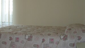 A bedroom with a bed covered in a patterned bedspread, a window with sheer curtains, and a white nightstand. - Villa Sandra (Giulianova)