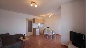 Ein Wohnzimmer mit Sofa, Fernseher, Esstisch mit Stühlen und einer Küchenzeile mit Schränken und Spüle.