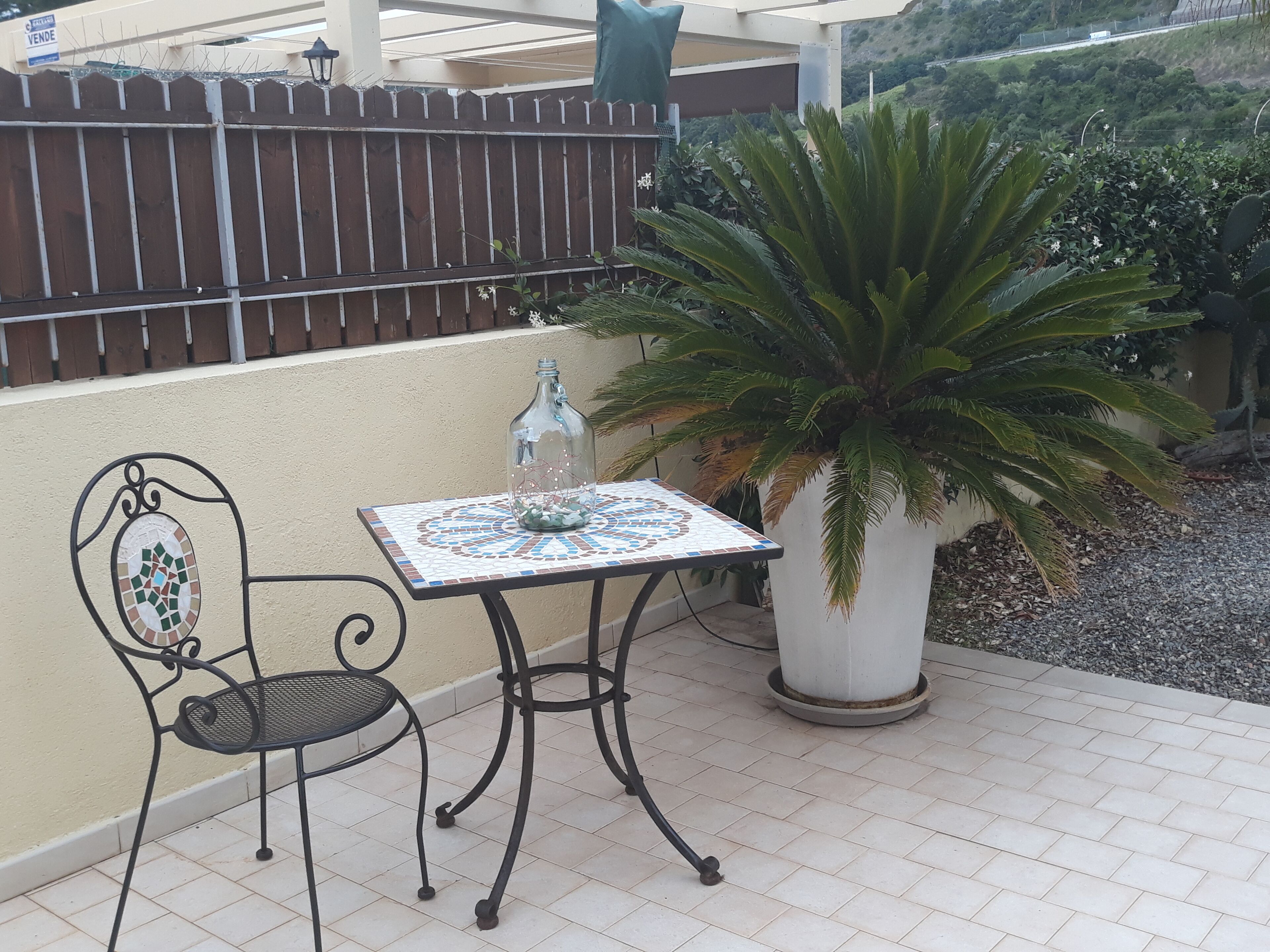 Une terrasse avec un sol carrelé, une chaise en métal, une table dont le plateau est décoré de mosaïques, une plante en pot et une clôture en arrière-plan.