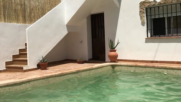 Una piscina amb vores de rajola, envoltada per una paret blanca amb una finestra i una porta. Hi ha escales que condueixen a la piscina, i diverses plantes en testos estan col·locades al voltant.