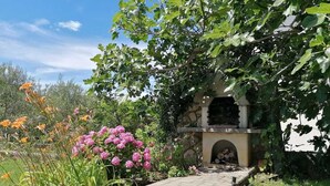 A stone pathway leading to a stone building with a fireplace, surrounded by lush greenery and flowering plants. - Apartment Grskovic G. Setaliste A2 (Silo - island Krk)
