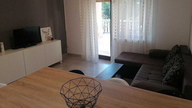A modern living room with a TV on a white cabinet, a wooden dining table, a sofa, and a view through a glass door. - Apartment Grskovic G. Setaliste A2 (Silo - island Krk)