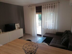 Una sala de estar moderna con un televisor sobre un mueble blanco, una mesa de comedor de madera, un sofá y vistas a través de un ventanal.