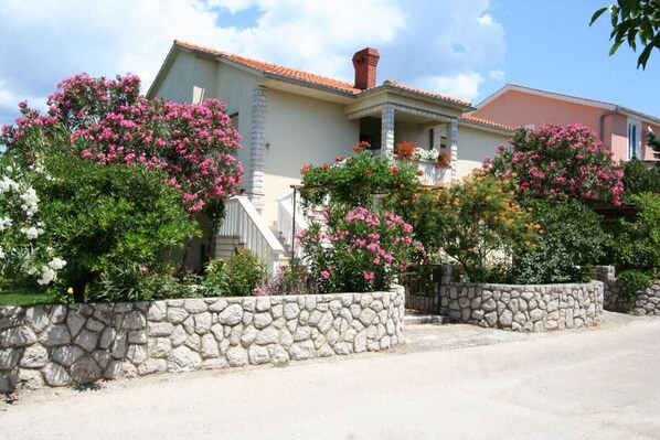 Una casa con un muro de piedra, arbustos florecientes de color rosa y un cielo despejado.