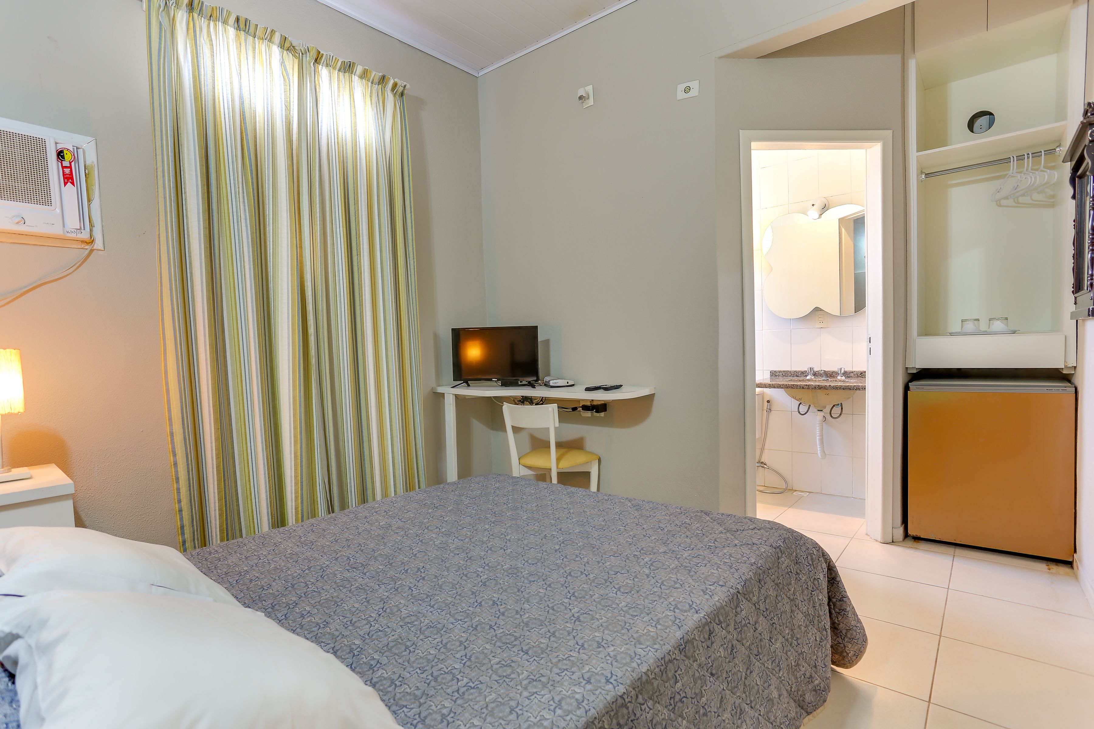 A hotel room with a bed, a desk with a computer, a small refrigerator, and a bathroom visible through an open door.