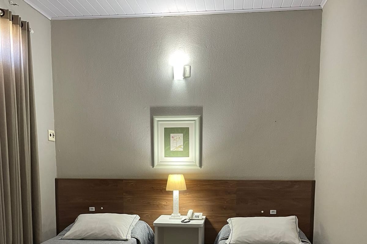 A hotel room with two single beds, a nightstand with a lamp, a framed picture on the wall, and a wooden headboard.