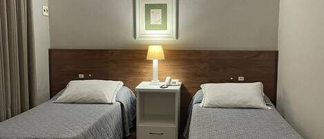 A hotel room with two single beds, a nightstand with a lamp, a framed picture on the wall, and a wooden headboard.