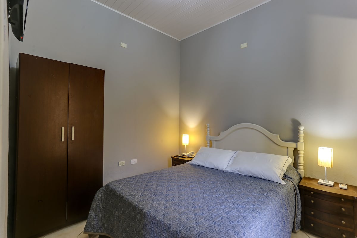 A bedroom with a bed, nightstands, a wooden wardrobe, and a wall-mounted TV.