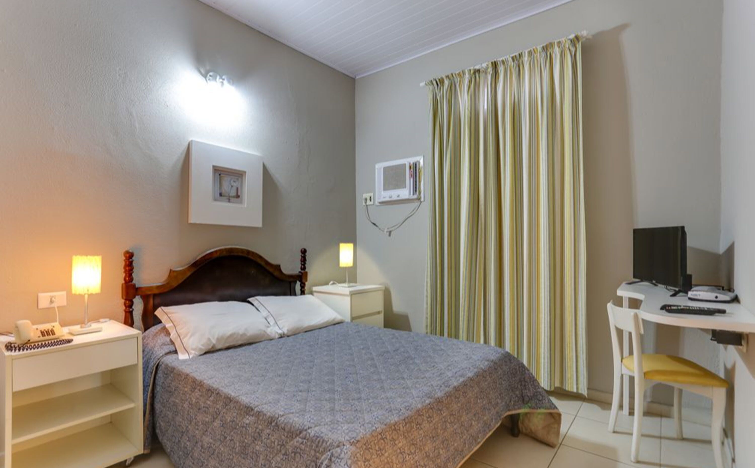 A hotel room with a bed, bedside tables, a desk with a chair, a TV, and a window with curtains.