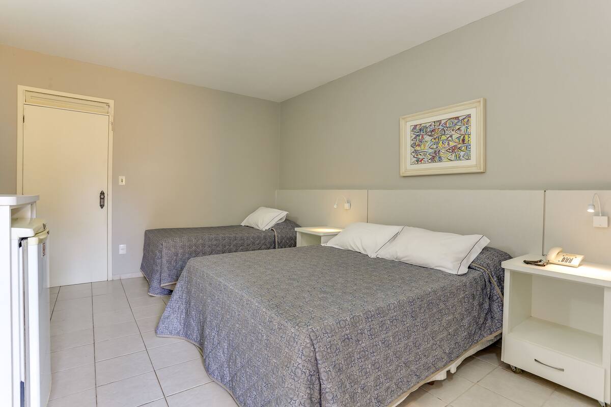 A room with two beds, a white bedside table with a lamp, a framed artwork on the wall, and a door.