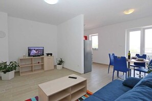 Uma sala de estar moderna com uma TV, uma área de refeições com cadeiras azuis e uma cozinha com geladeira.