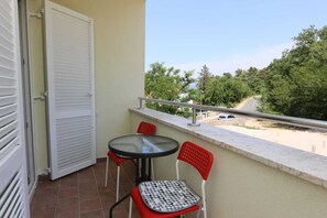 Varanda com mesa e duas cadeiras, uma porta branca e vista para árvores e uma estrada.