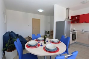 Uma área de jantar com uma mesa redonda posta para quatro pessoas, cadeiras azuis, uma garrafa de vinho e um vaso de planta.