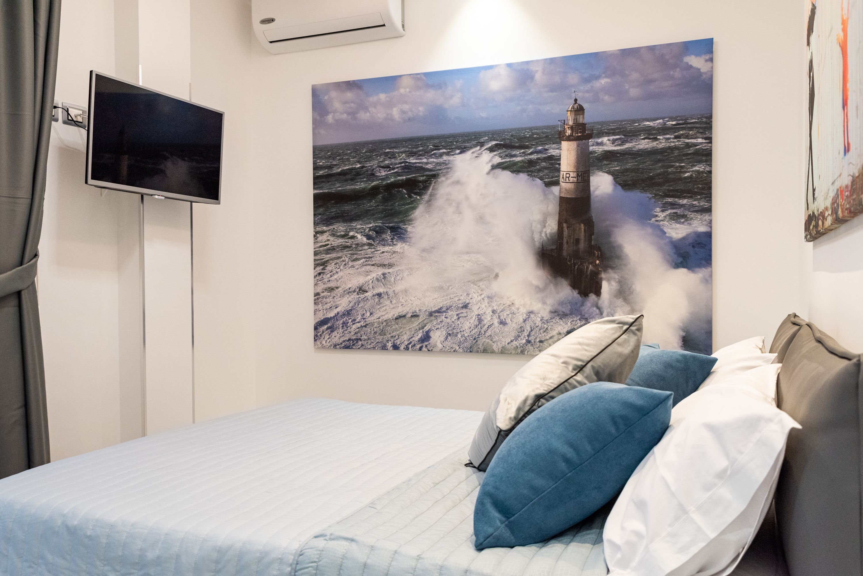 Una camera da letto con un grande quadro raffigurante un faro, una TV a schermo piatto montata a parete e un letto con lenzuola bianche e azzurre.