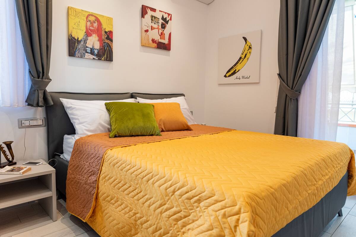 Una cama bien hecha con colcha amarilla, una mesita de noche con lámpara y cuadros en las paredes.