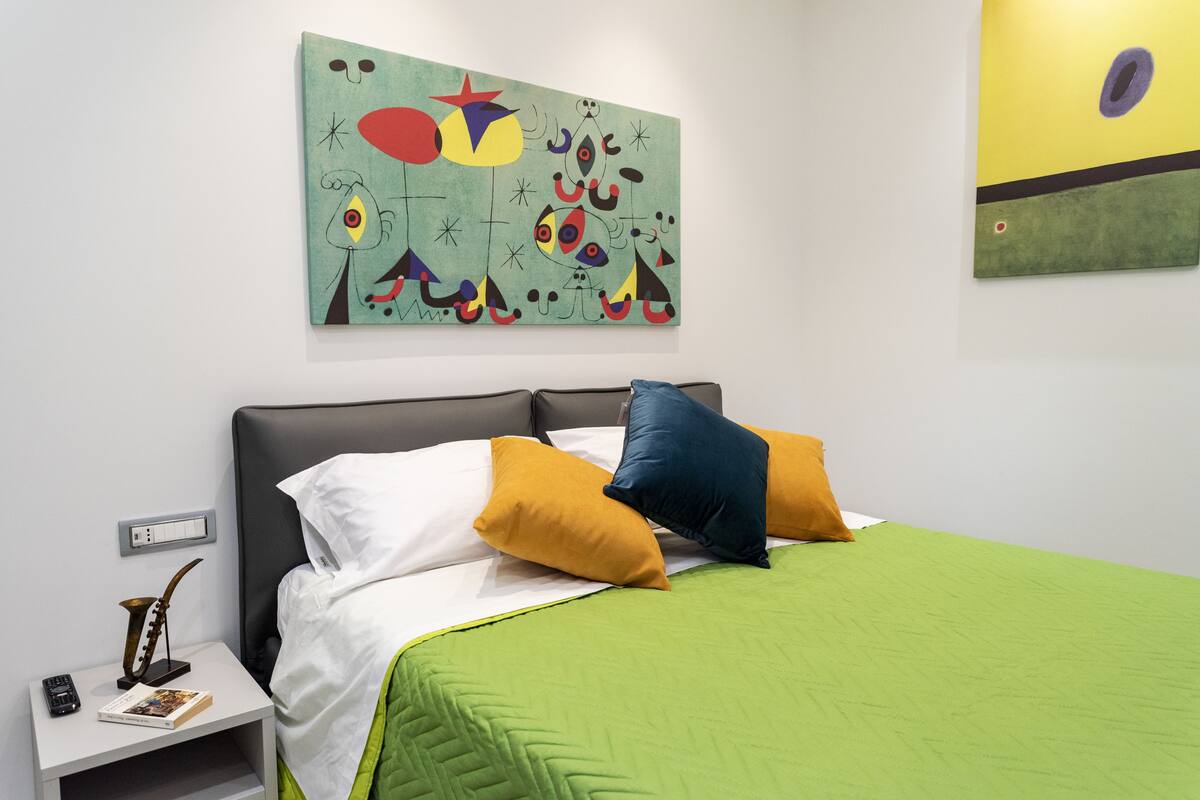 Un dormitorio con cama, almohadas, una mesita de noche, lámpara y arte abstracto en las paredes.