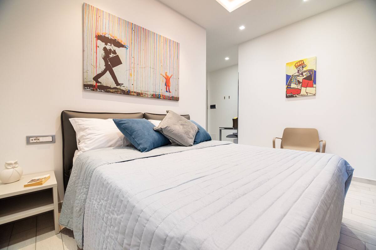 Un dormitorio moderno con una cama grande, una mesita de noche con un libro y dos obras de arte colgadas en la pared.