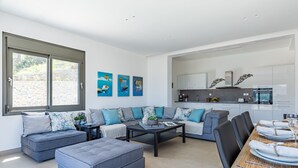 Ein moderner Wohnraum mit einer Sofagarnitur, Hocker, Couchtisch und einem Essbereich mit Holztisch und Stühlen.