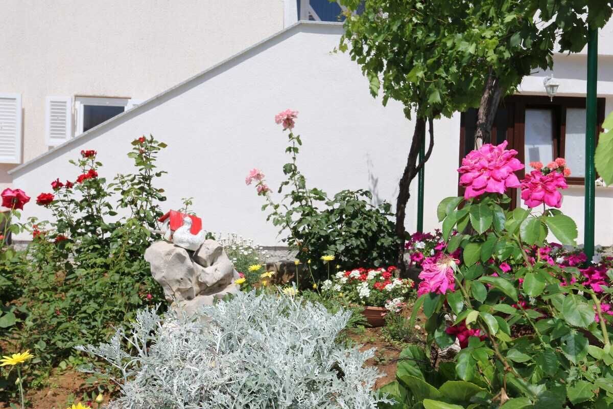 En trädgård med olika växter och blommor, en stenskulptur och en byggnad med fönster i bakgrunden.