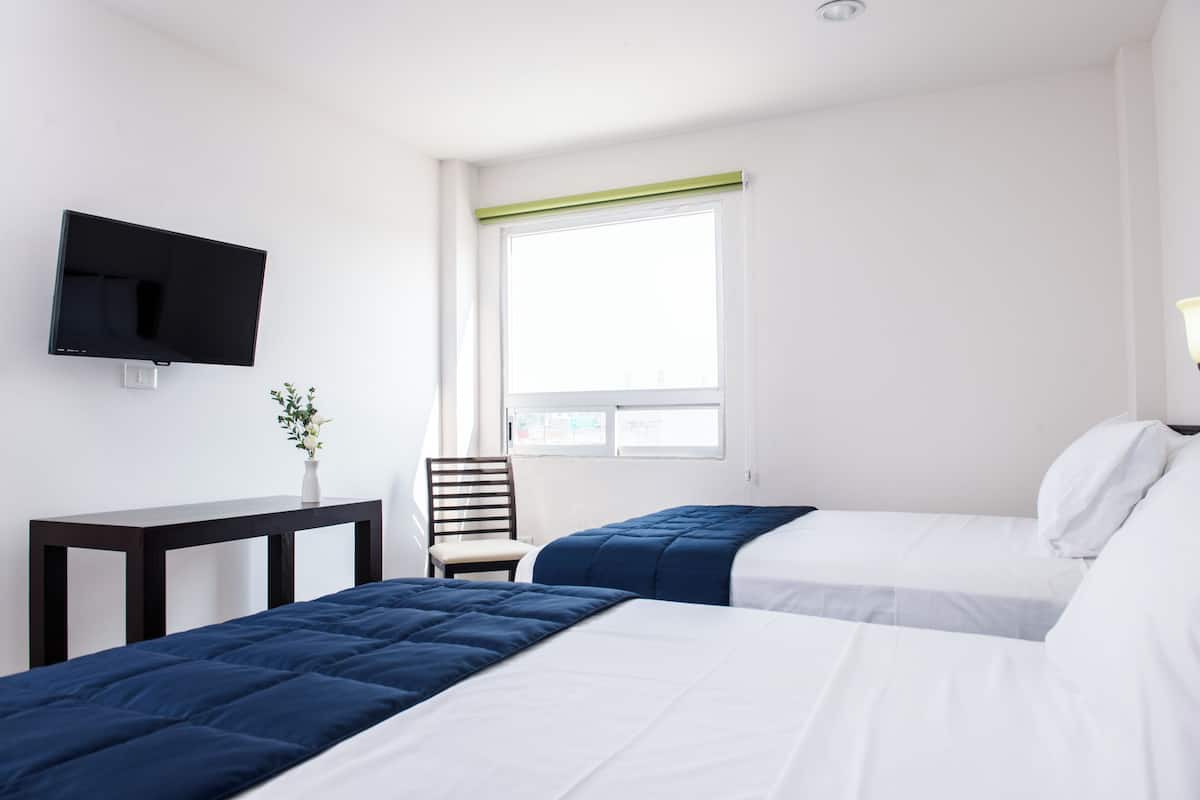 Ein Hotelzimmer mit zwei Betten, einem an der Wand befestigten Fernseher, einem kleinen Tisch mit einer Vase und einem Fenster mit Blick.