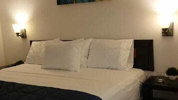 Habitación de hotel con una cama grande, ropa de cama blanca, una manta azul, dos mesitas de noche con lámparas y un cuadro abstracto y colorido en la pared.