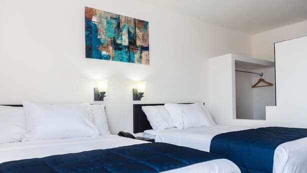 Une chambre d’hôtel avec deux lits, du linge de lit blanc, une couvre-lit bleue, un tableau au mur et un porte-manteau en bois sur la porte.