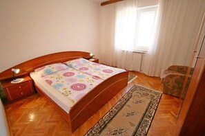 Een slaapkamer met een houten bed, nachtkastje, stoel, raam met gordijnen en een kamerbreed tapijt met patroon.