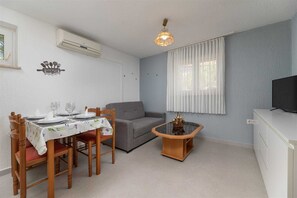 Ein Essbereich mit einem Tisch für vier Personen, einem grauen Sofa, einem kleinen Holztischchen mit einer Vase und einem Fernseher auf einem Schrank.
