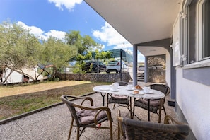 Une terrasse extérieure avec des chaises et une table en osier, un mur de pierre et une vue sur les voitures garées et les arbres.