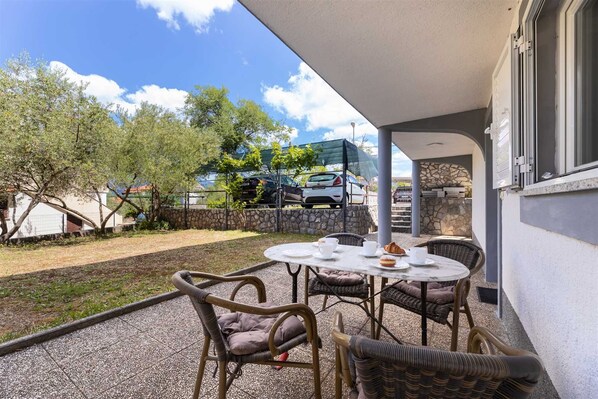 An outdoor patio with wicker chairs and a table, a stone wall, and a view of parked cars and trees.