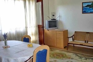 Une pièce comprenant une table à manger, des chaises, un téléviseur posé sur un meuble en bois, un canapé et un tableau accroché au mur.