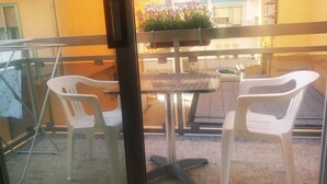 Een balkon met twee witte kunststof stoelen en een klein tafeltje, een raam en uitzicht op een gebouw met bloemen.